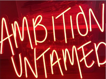 Red neon sign spelling out “AMBITION UNTAMED” in a bold all-caps font, glowing brightly against a dark background.