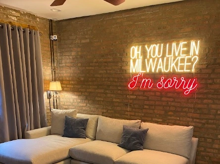 Custom neon sign in white and red text reading “Oh, you live in Milwaukee? I’m sorry,” glowing against a brick wall above a cozy beige sectional sofa.